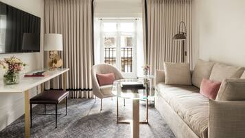 A hotel room with a sofa, a glass coffee table, a chair, a desk, a TV, and a window with curtains.