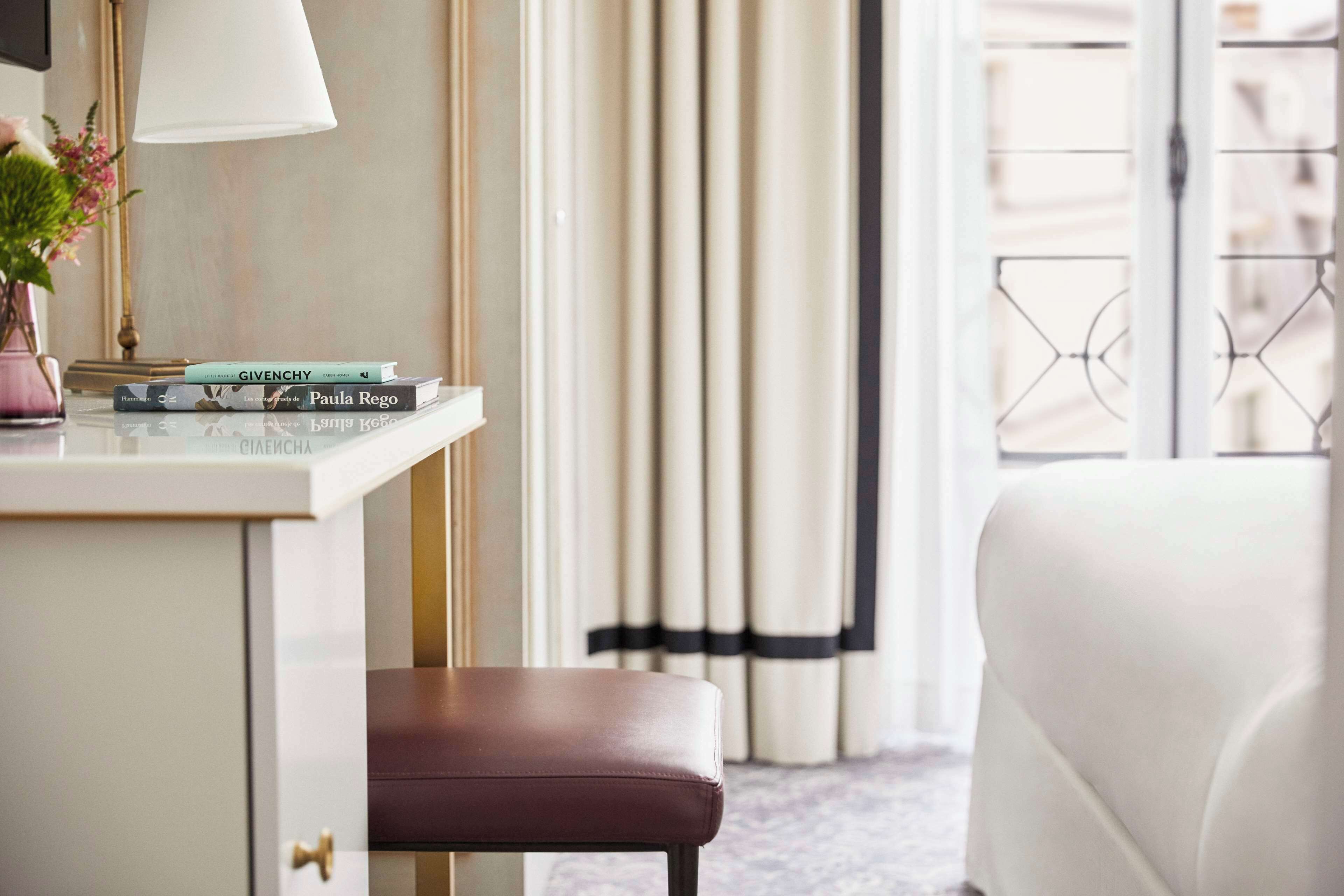 A hotel room with a white desk, a brown leather bench, a white sofa, and a window with curtains.