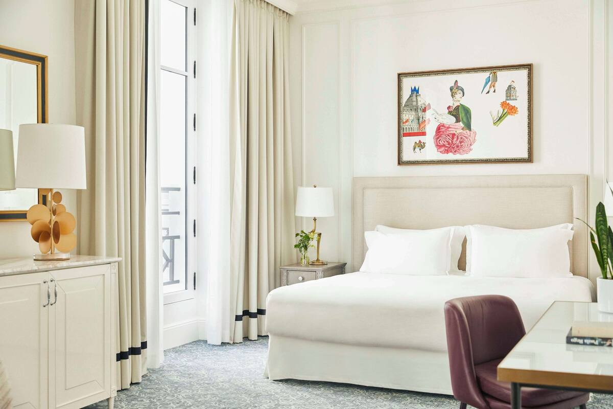 A hotel room with a large bed, a desk, a chair, a nightstand with a lamp, a mirror, and a wall with a framed picture.