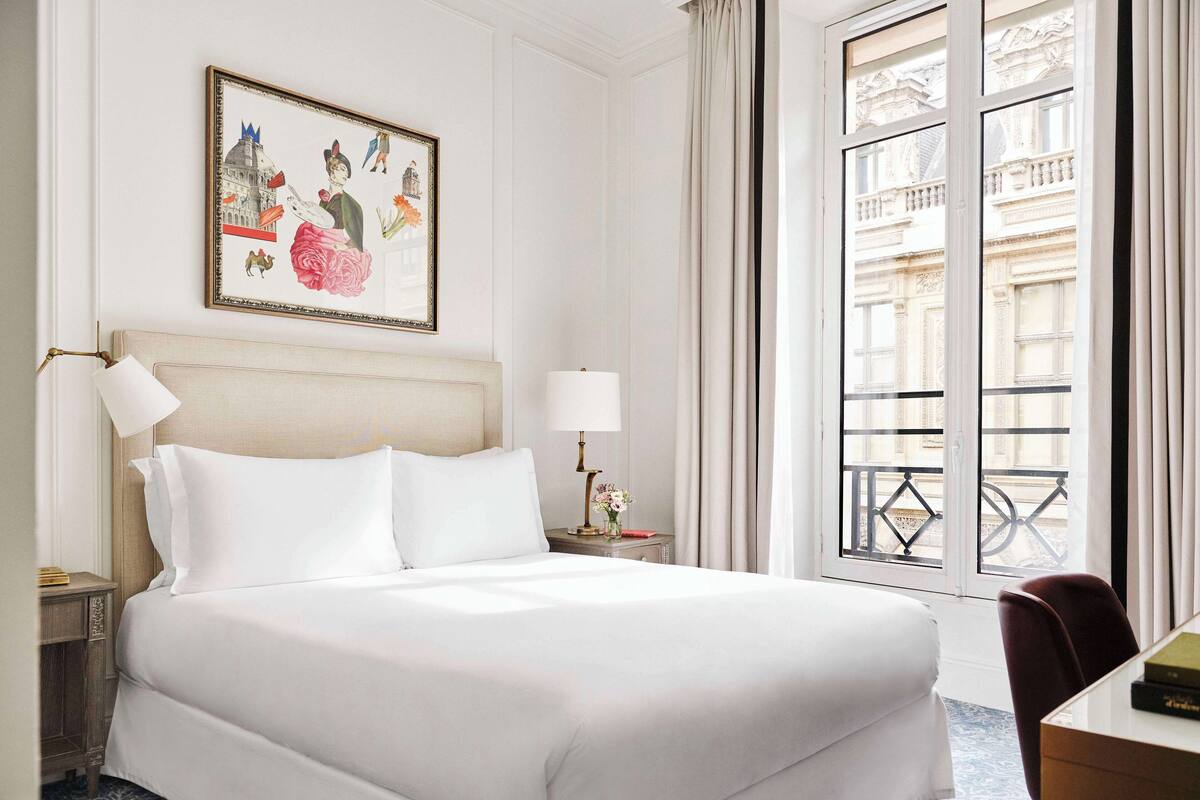 A hotel room with a large bed, a nightstand with a lamp, a framed picture on the wall, and a view of a building through the window.