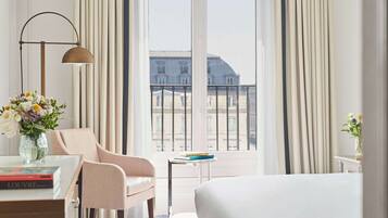 A hotel room with a large bed, a desk with a lamp, a chair, a small table with books, and a view of the city through large windows.