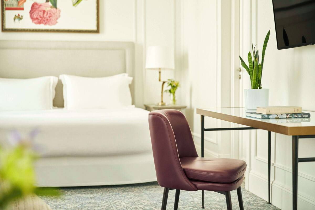 A hotel room with a bed, a desk with a chair, a potted plant, and a television on the wall.
