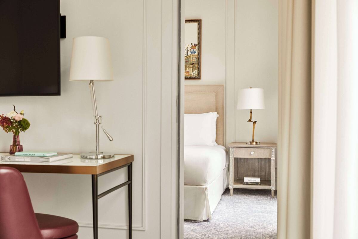 A hotel room with a bed, bedside table, lamp, desk, chair, and a small plant.