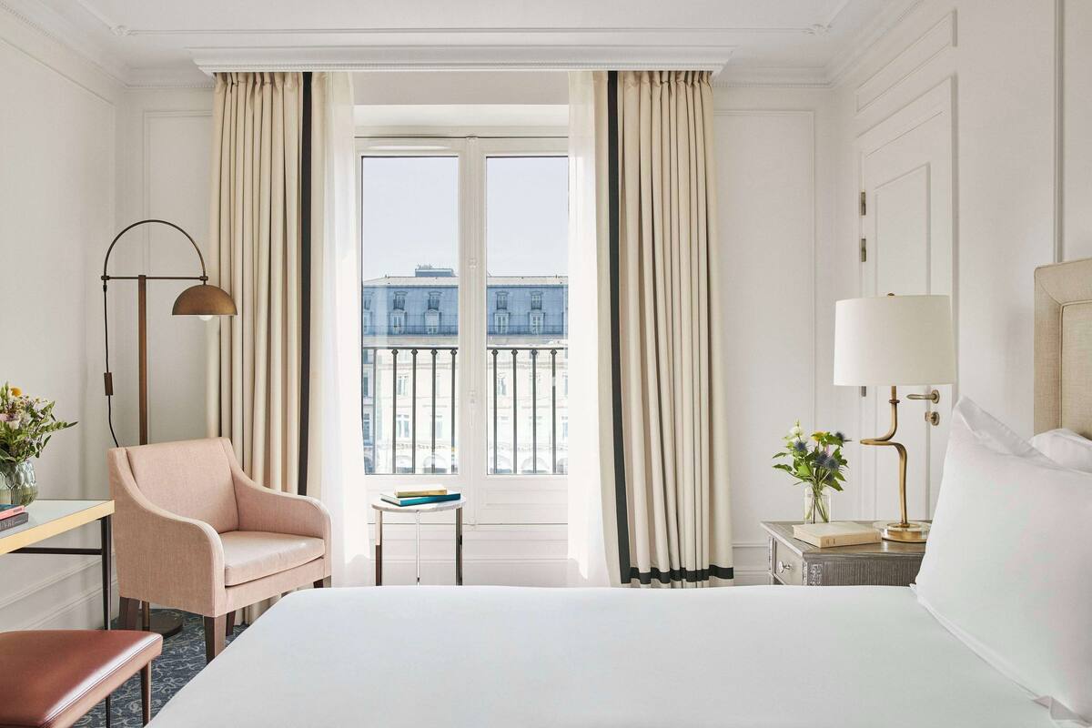 A hotel room with a large bed, a desk with a chair, a bedside table with a lamp, and a view of the city through large windows.