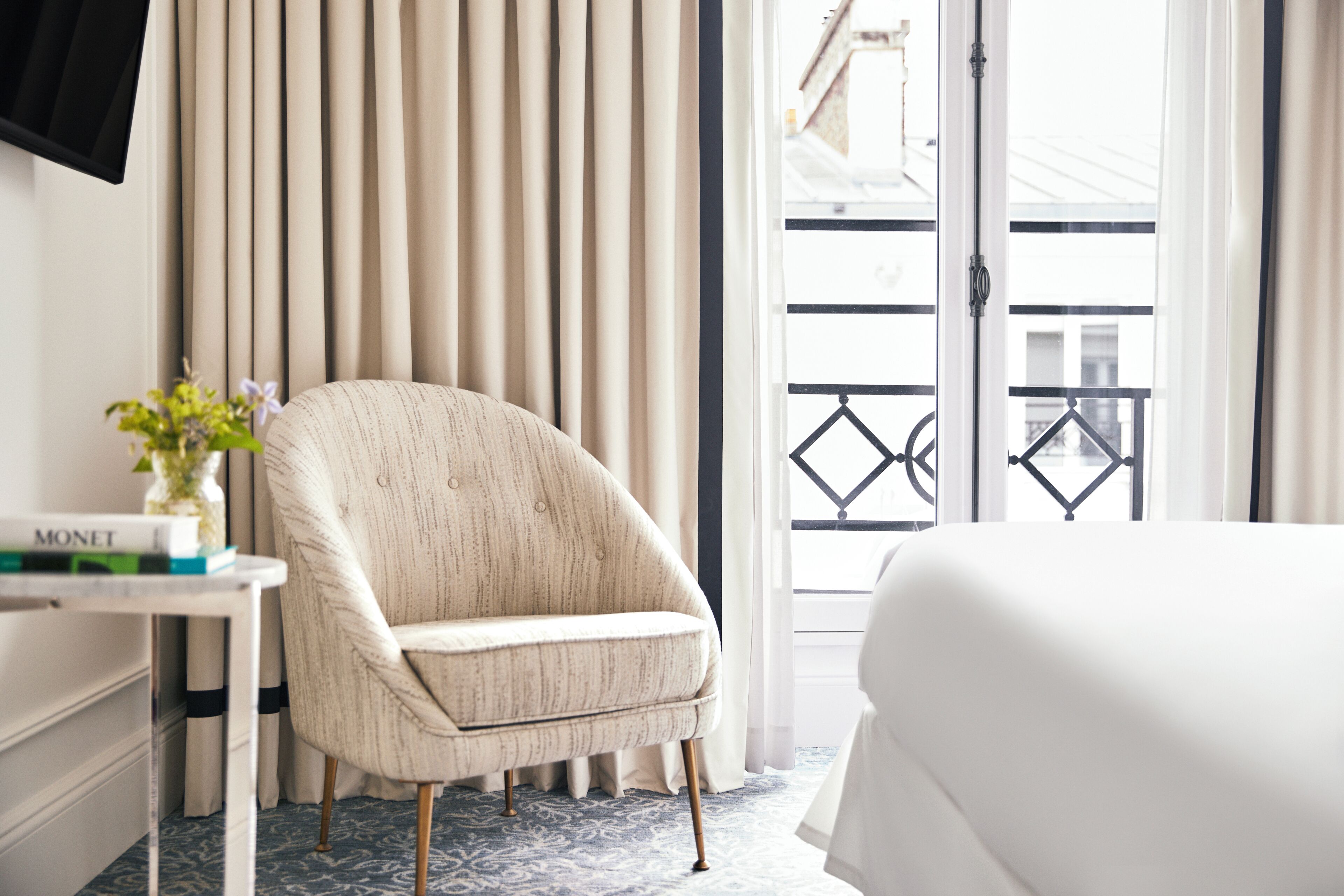 Habitación de hotel con cama, una silla, un escritorio con un florero y vistas al exterior a través de una ventana con cortinas.