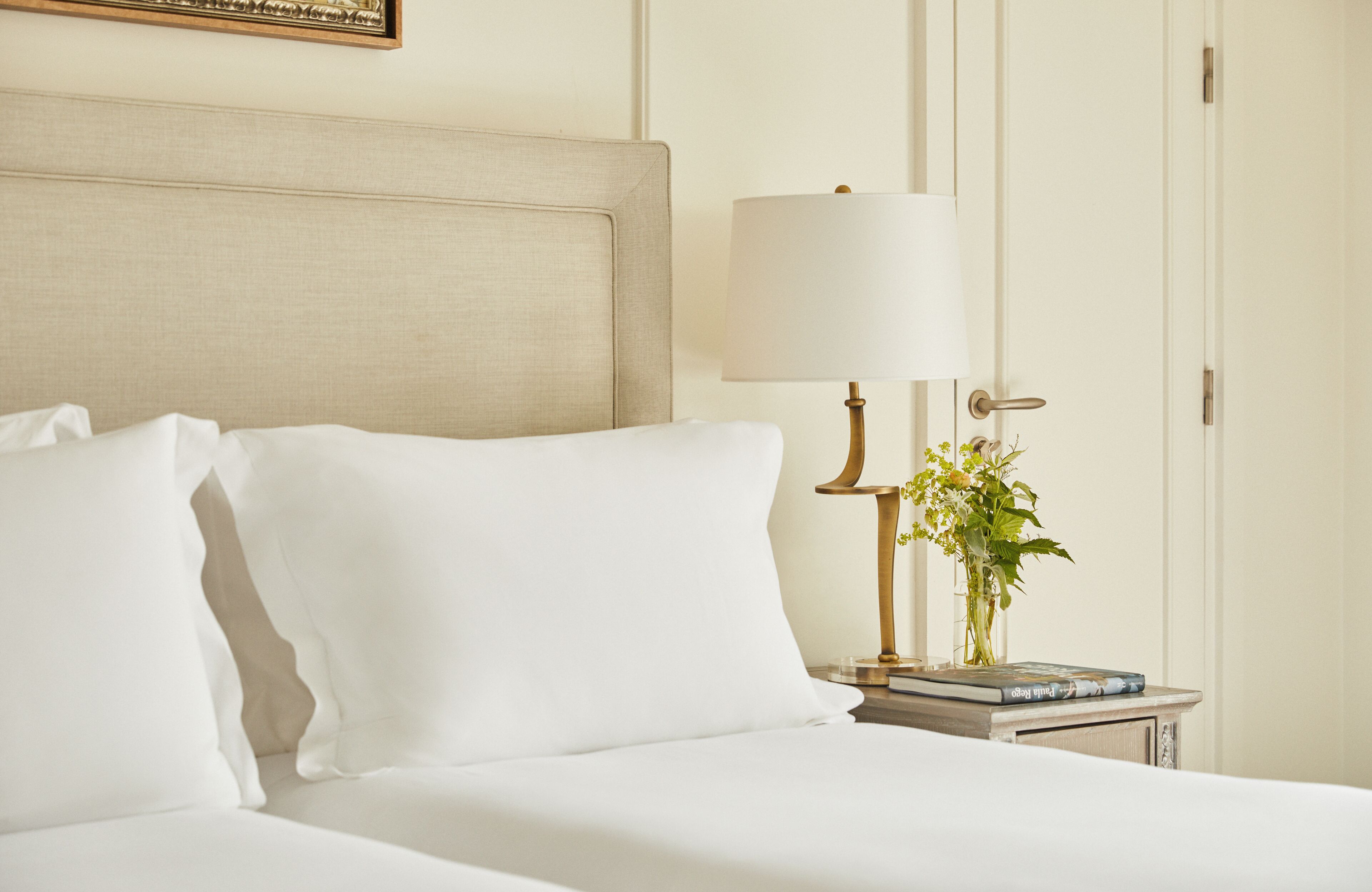A neatly made bed with white linens, a bedside lamp, and a vase with flowers on a nightstand.