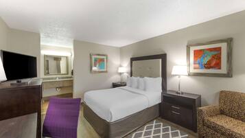 A hotel room with a bed, a TV, a desk, a chair, a lamp, and artwork on the walls.