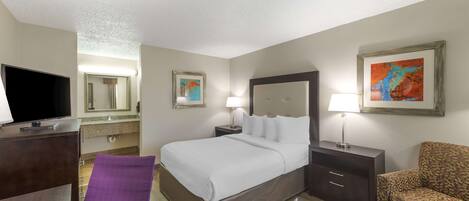 A hotel room with a bed, a TV, a desk, a chair, a lamp, and artwork on the walls.