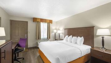 A hotel room with a large bed, a desk with a chair, a nightstand with a lamp, and a door.