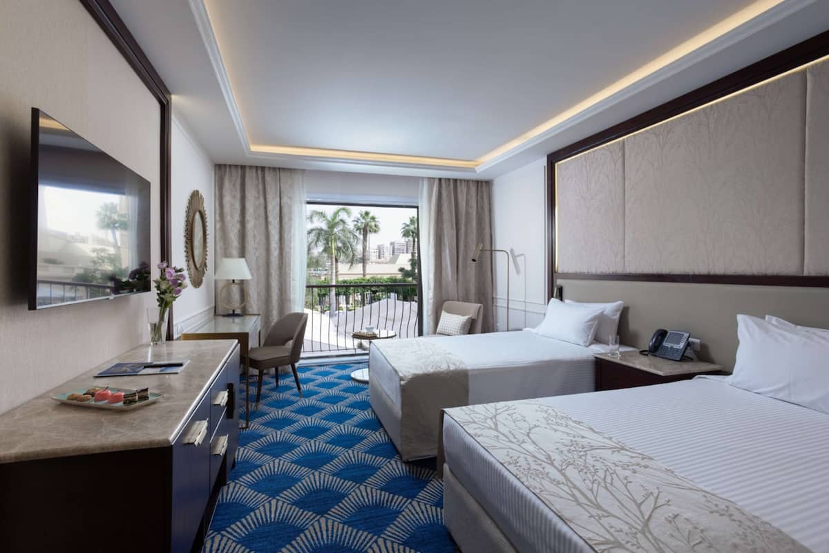 A hotel room with two beds, a desk with a chair, a TV, and a balcony with a view of palm trees and buildings.