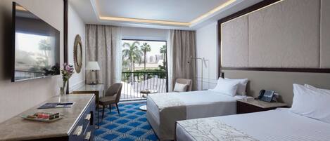 A hotel room with two beds, a desk with a chair, a TV, and a balcony with a view of palm trees and buildings.