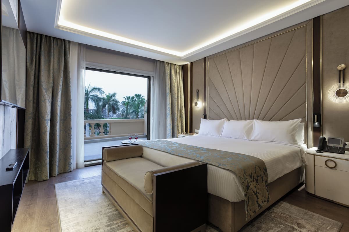 A modern hotel room with a large bed, a sofa, a bedside table, and a window with a view of palm trees.