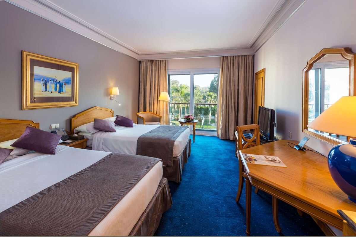 A hotel room with two beds, a desk, a chair, a TV, and a balcony with curtains.