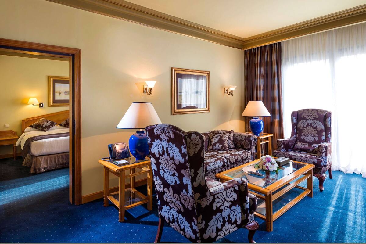 A hotel room with a bed, a sofa, two armchairs, a coffee table, and a desk with a phone.