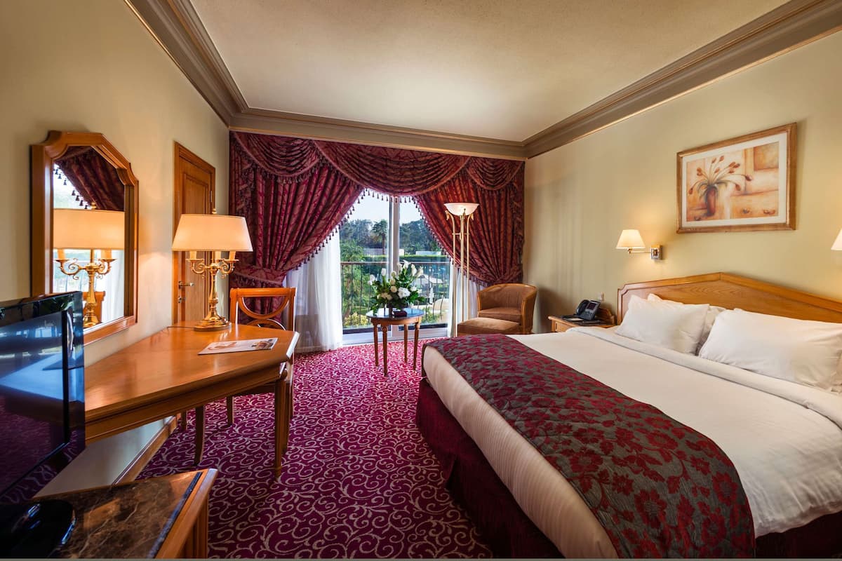 A hotel room with a large bed, a desk with a lamp, a chair, a TV, and a balcony with a view.