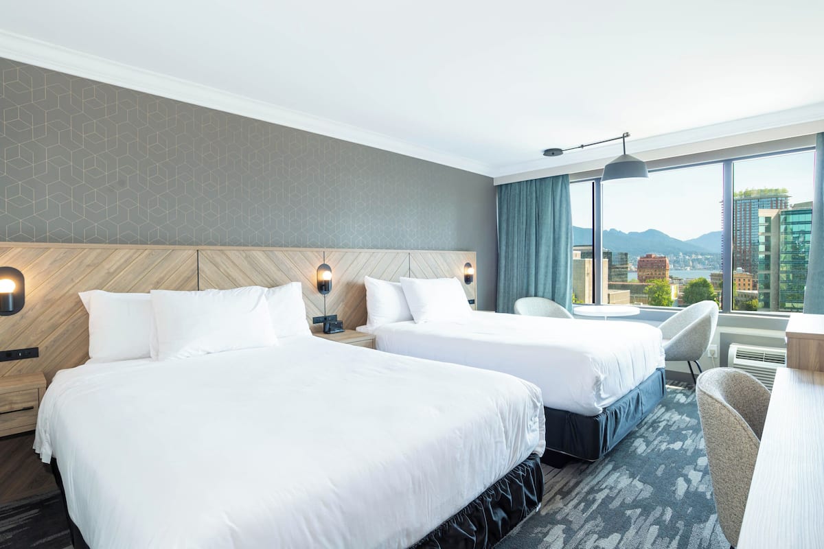 Een hotelkamer met twee bedden, een bureau, een stoel en een groot raam met uitzicht op de stad.