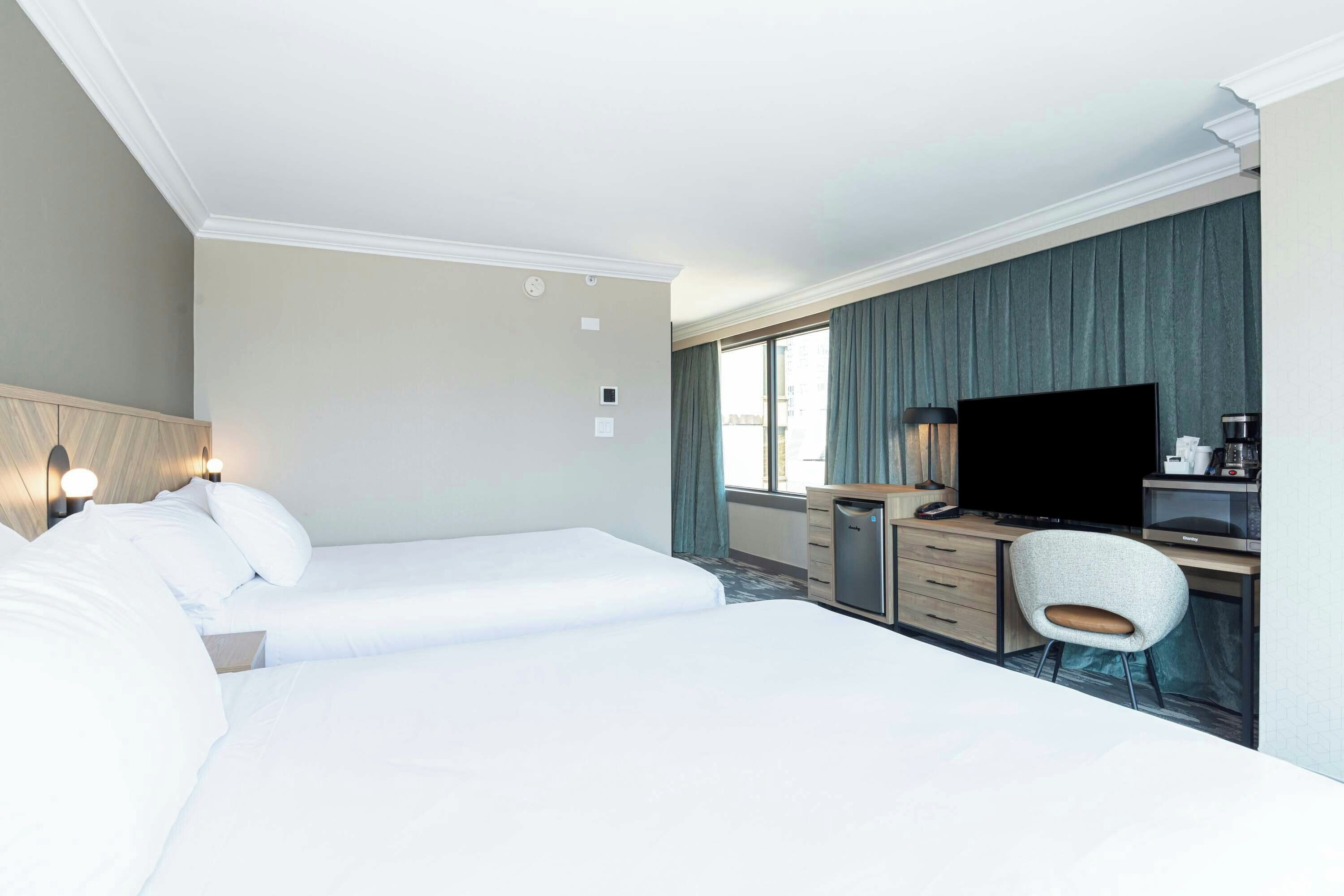 A hotel room with two beds, a desk with a flat-screen TV, a chair, and a small kitchenette.