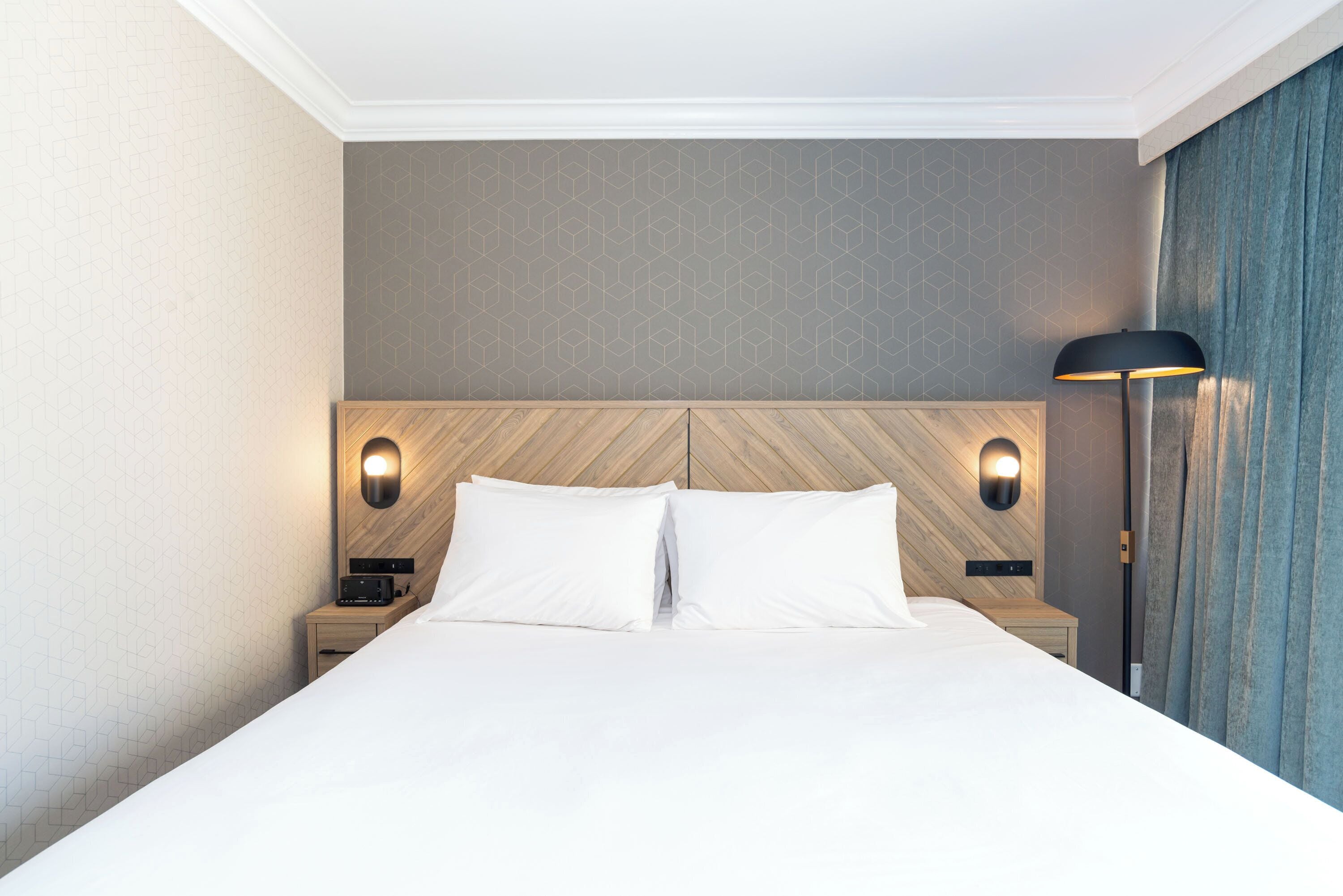 A hotel room with a large bed, two bedside tables, a nightstand, a wall-mounted lamp, and a curtain.