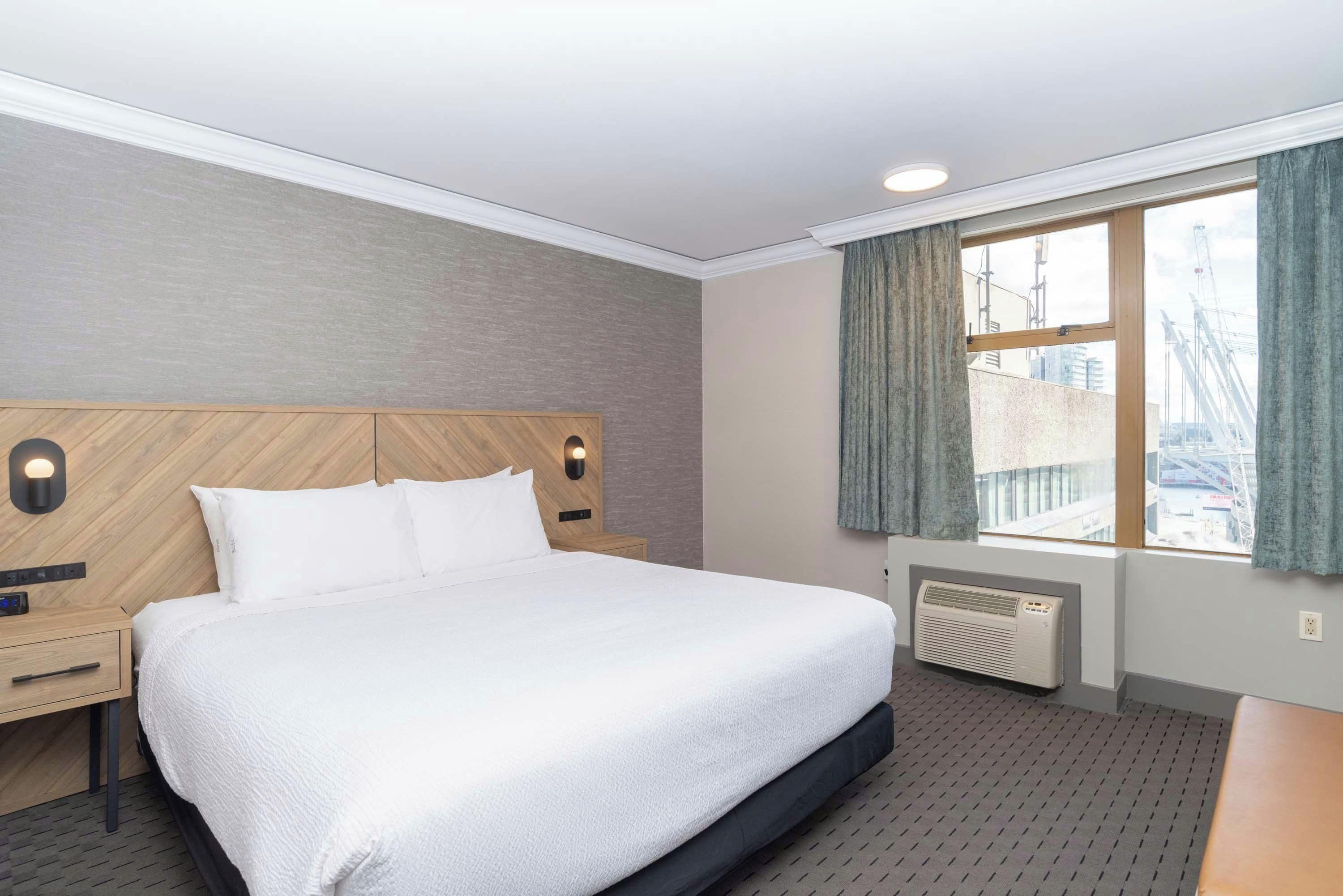 Une chambre d’hôtel comprenant un lit, une table de chevet, une lampe fixée au mur, une fenêtre avec des rideaux et un radiateur.