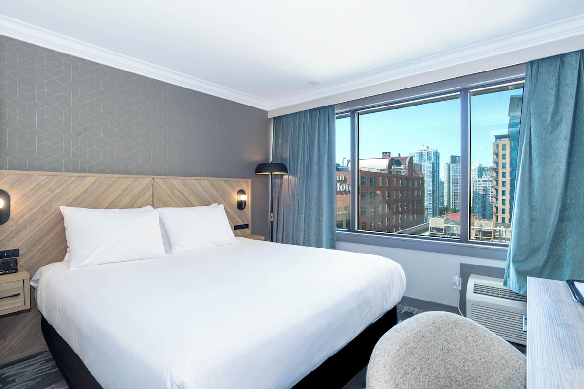 Hotelkamer met een groot bed, nachtkastje, lamp en uitzicht op de stad door een groot raam.