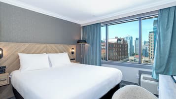 A hotel room with a large bed, a nightstand, a lamp, a window with curtains, and a view of the cityscape.