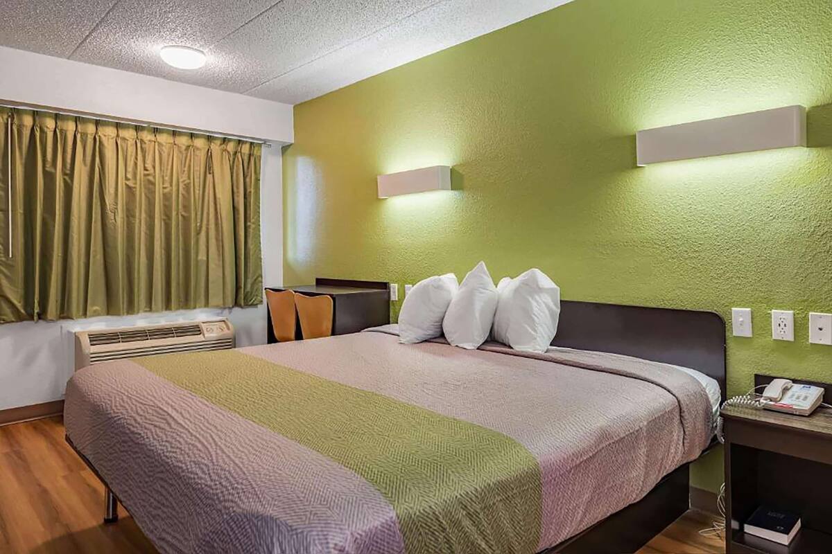A hotel room with a large bed, a bedside table with a phone, a wall-mounted light, and a window with curtains.