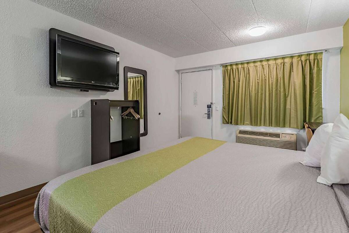 A hotel room with a bed, a television mounted on the wall, a mirror, a coat rack, and a window with curtains.