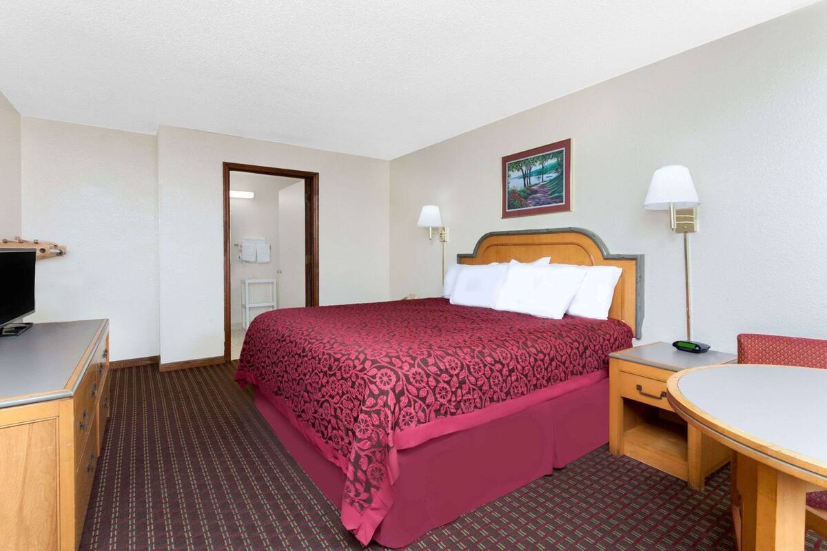 A hotel room with a bed, a desk, a chair, a TV, a painting on the wall, and a door.