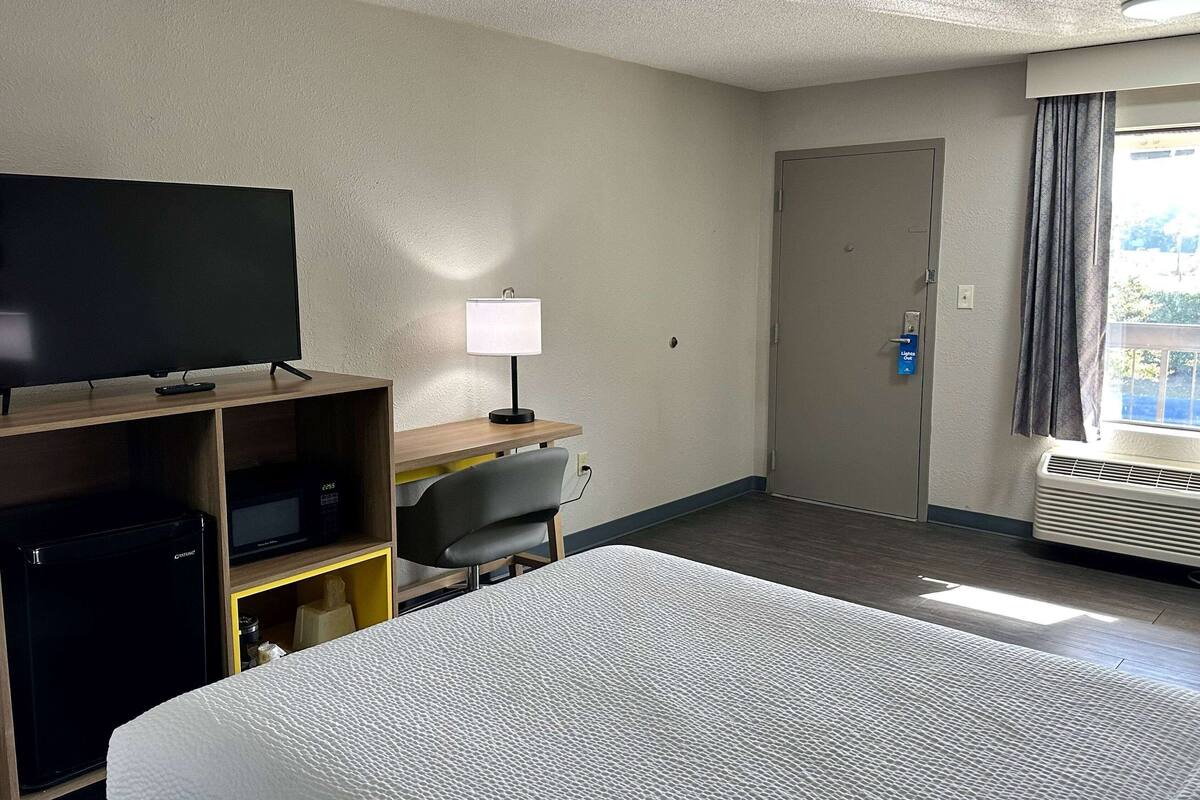 A hotel room with a bed, a desk with a lamp, a television, a mini-fridge, and a door.