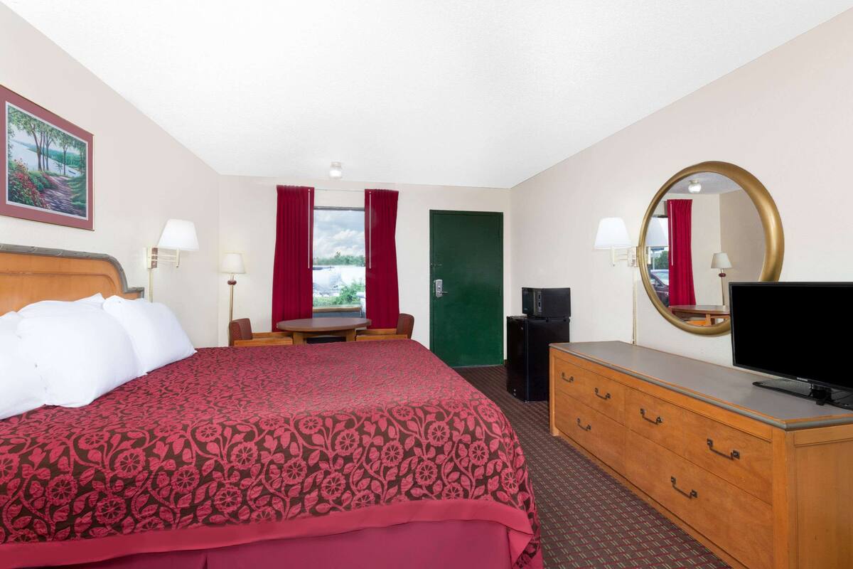 A hotel room with a large bed, a dresser, a television, and a window with red curtains.
