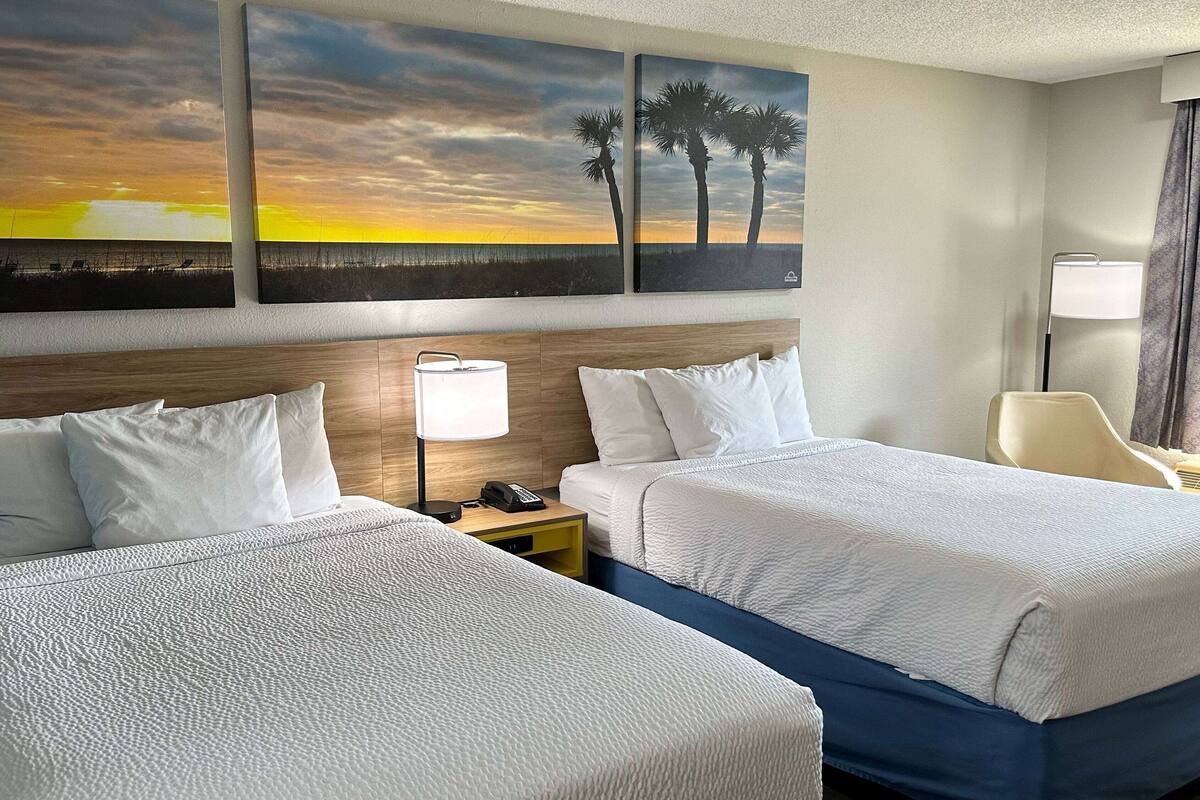 A hotel room with two beds, a wooden headboard, a nightstand with a lamp, and artwork depicting a sunset and palm trees.