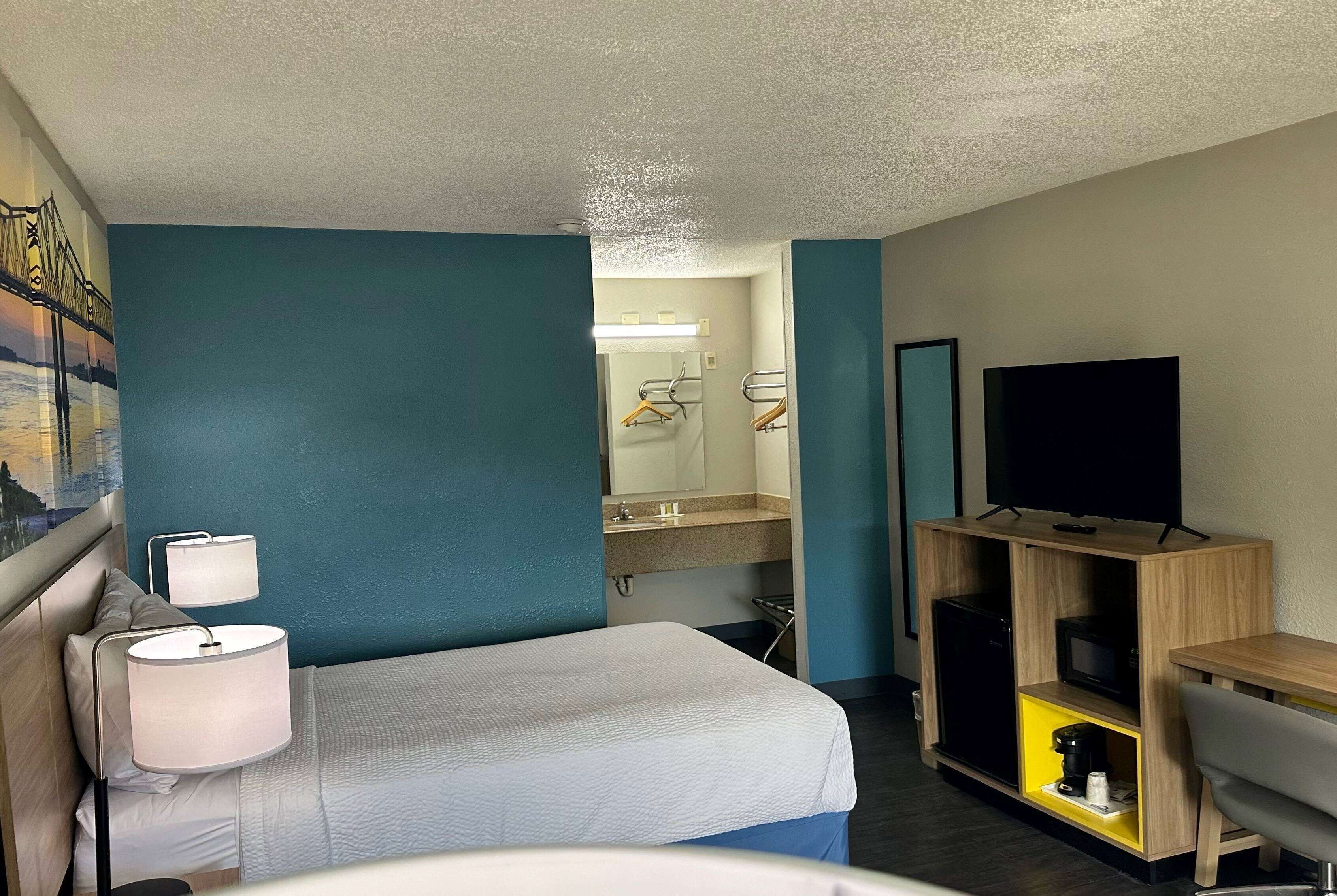 A hotel room with a bed, a desk, a TV, a mirror, and a bathroom visible through an open door.
