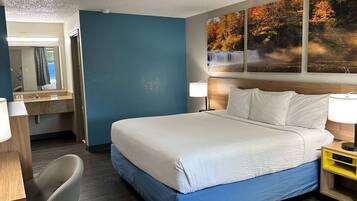 Habitación de hotel con una cama grande, un escritorio con una silla, un baño con espejo y un cuadro de temática natural en la pared.