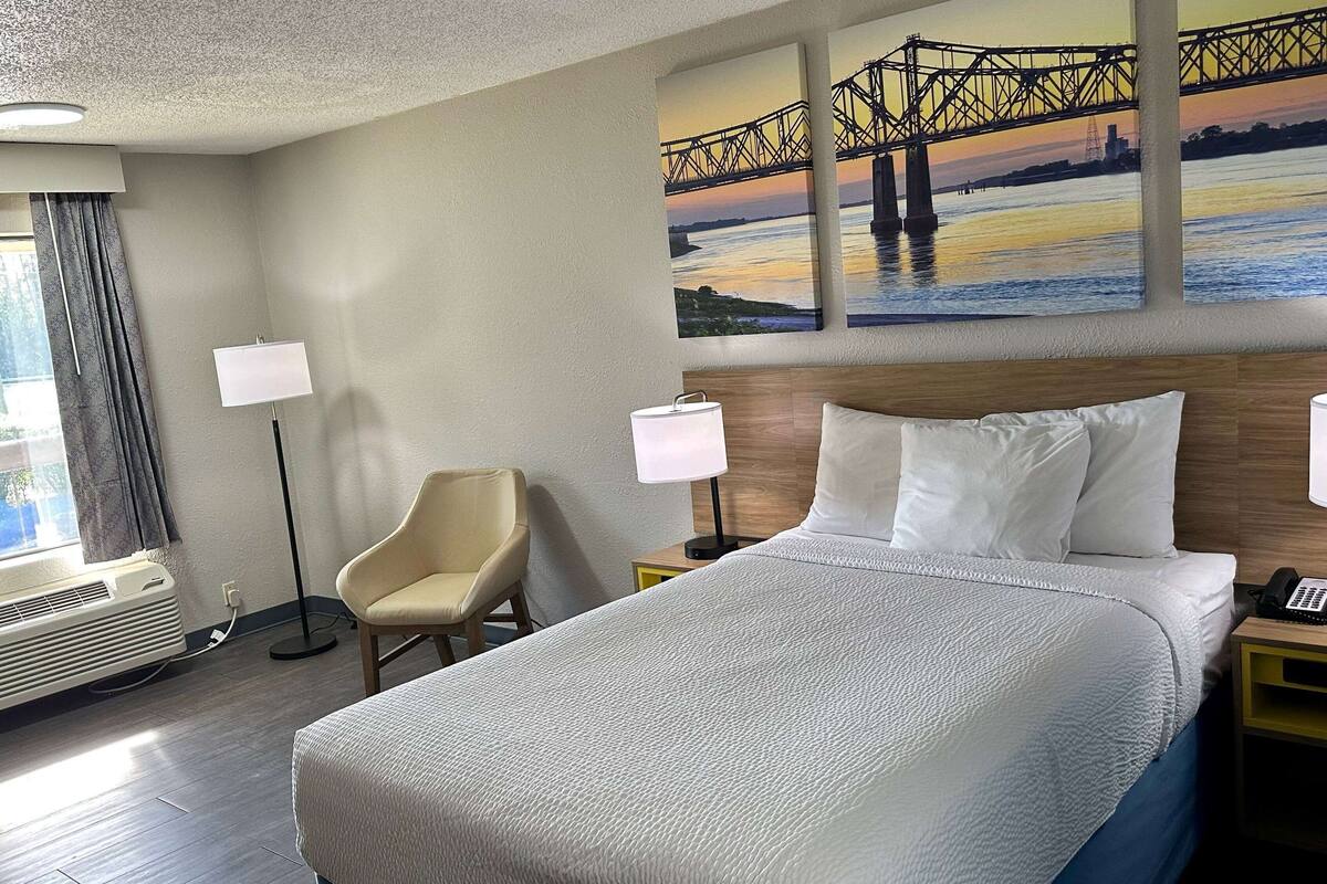 A hotel room with a large bed, two bedside lamps, a chair, and a painting of a bridge on the wall.