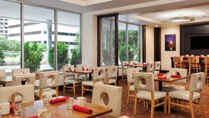 Un comedor de hotel con varias mesas y sillas, ventanas amplias y una pantalla de televisión.