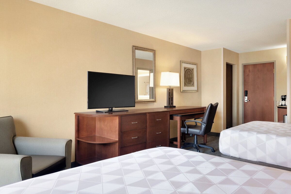 Habitación de hotel con dos camas, un escritorio con televisor, una silla, una lámpara y una puerta.