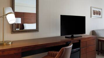 Quarto de hotel com escrivaninha de madeira, televisão, espelho, cadeira e mesa pequena.