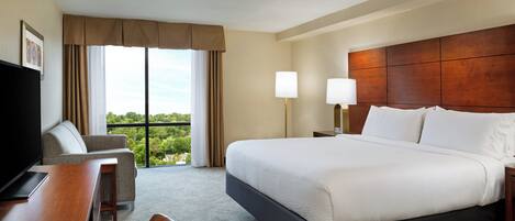 A hotel room with a large bed, a desk with a chair, a television, and a view of greenery through the window.