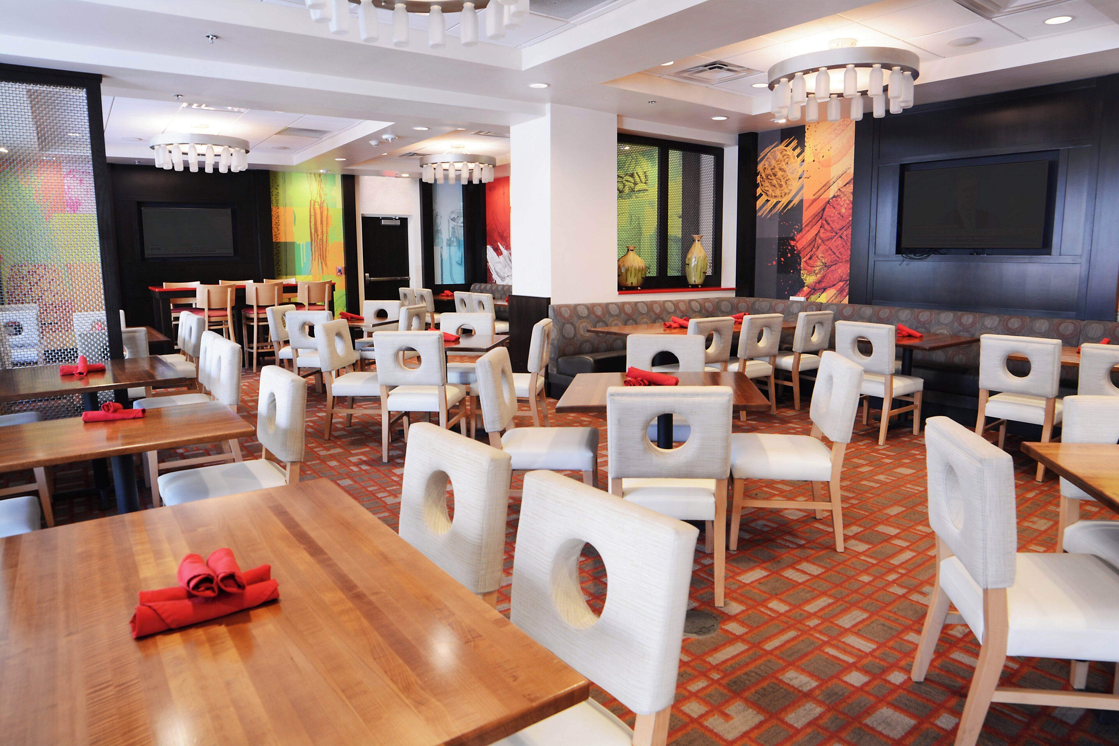 A restaurant with multiple tables and chairs, a patterned carpet, and a large screen on the wall.