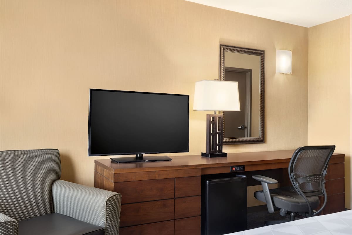 A hotel room with a flat-screen TV, a desk with a lamp, a chair, and a bed.
