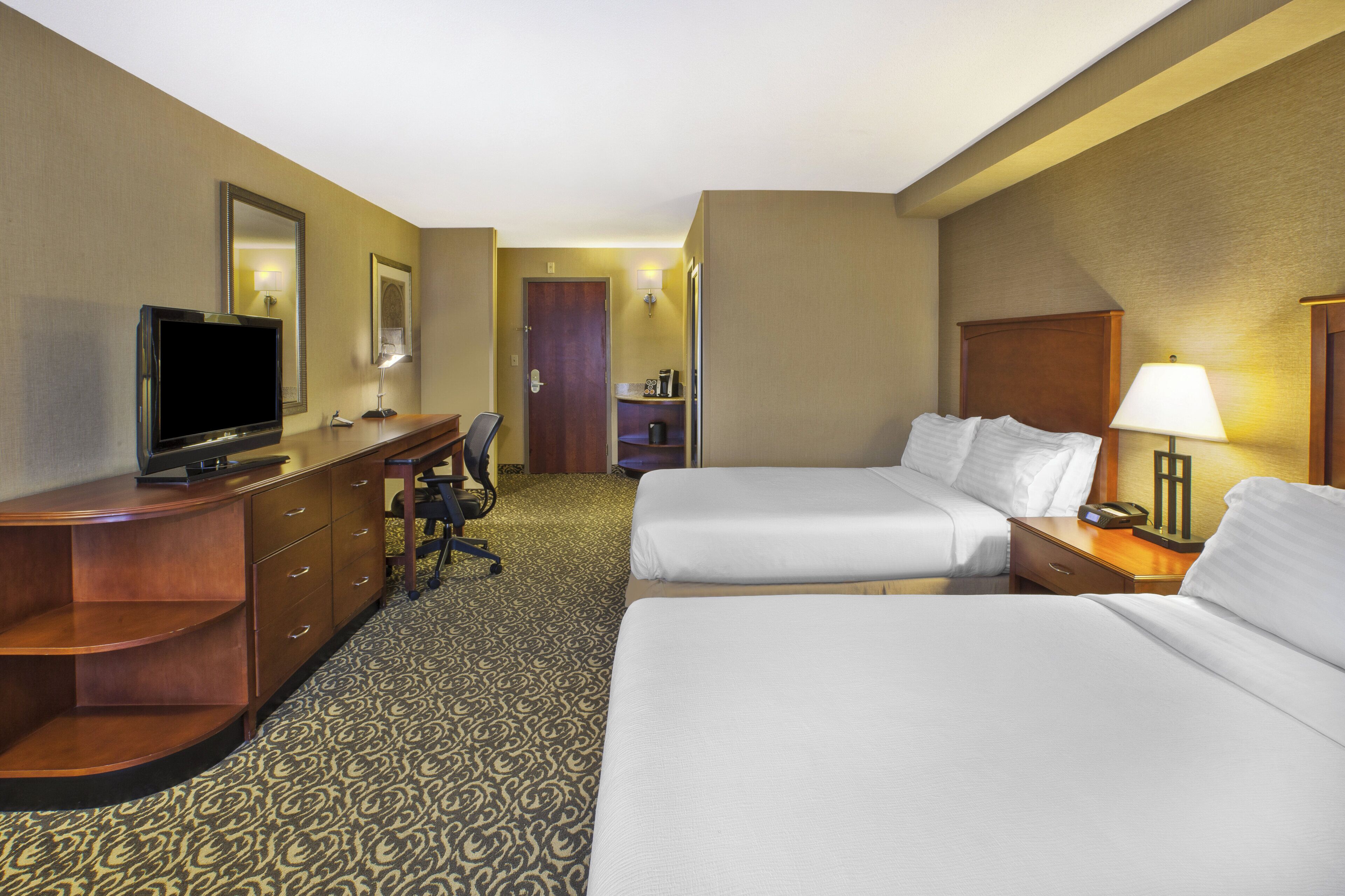A hotel room with two beds, a desk with a television, a chair, and a lamp.