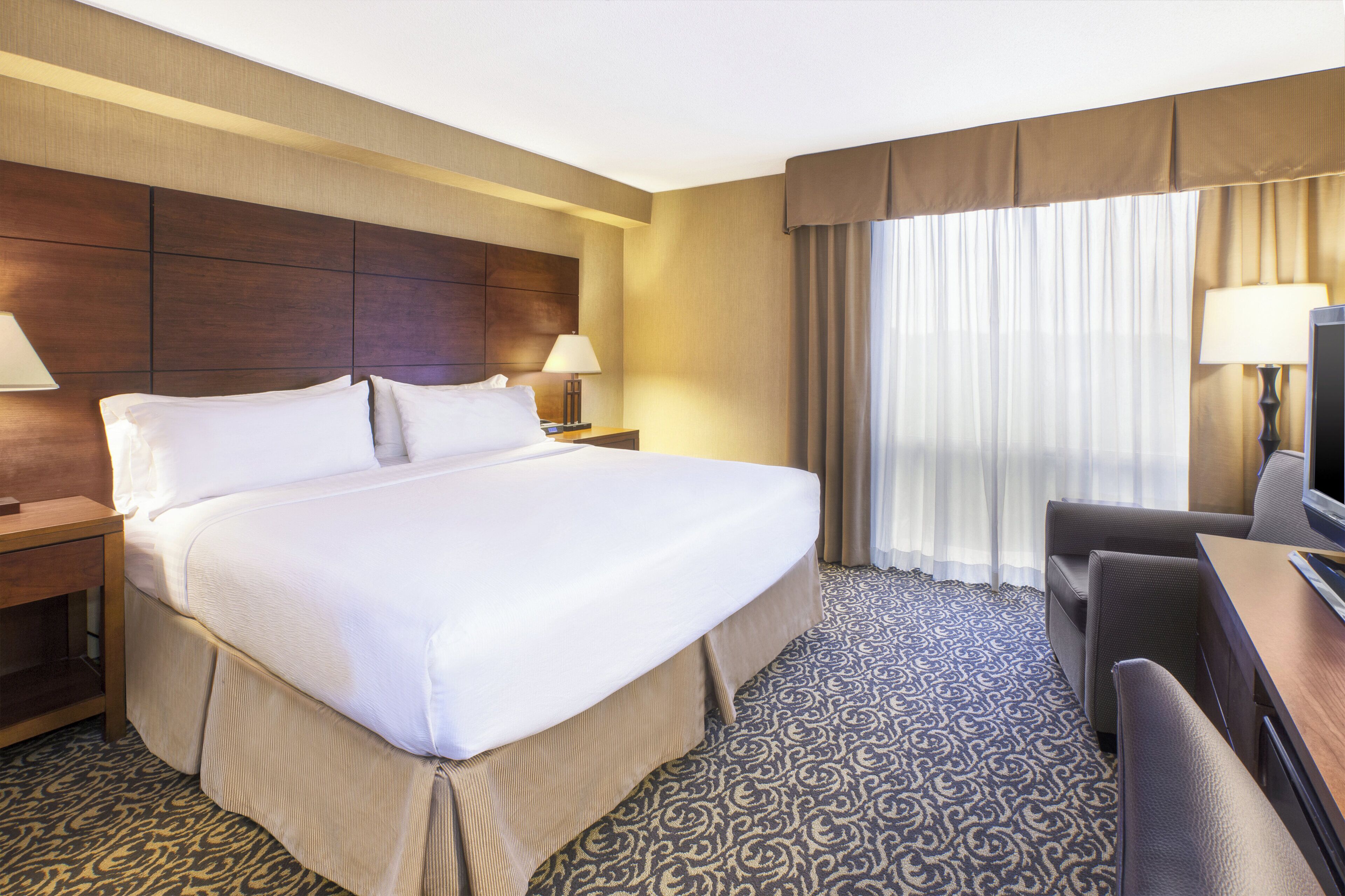 A hotel room with a large bed, two bedside tables, a desk with a chair, a television, and a window with curtains.