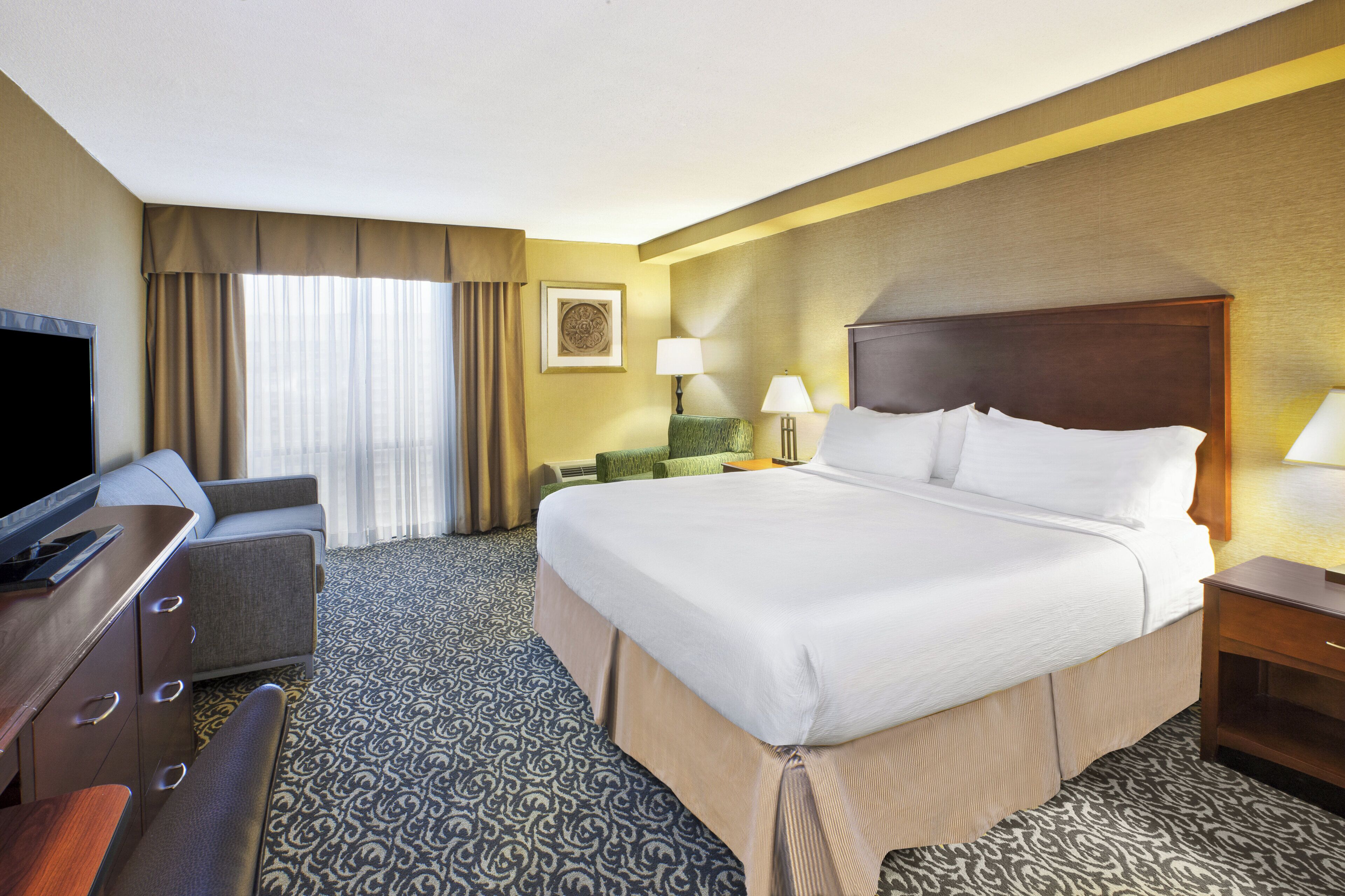 Quarto de hotel com cama, escrivaninha, televisão, luminária e janela com cortinas.