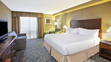 Quarto de hotel com cama, escrivaninha, televisão, luminária e janela com cortinas.
