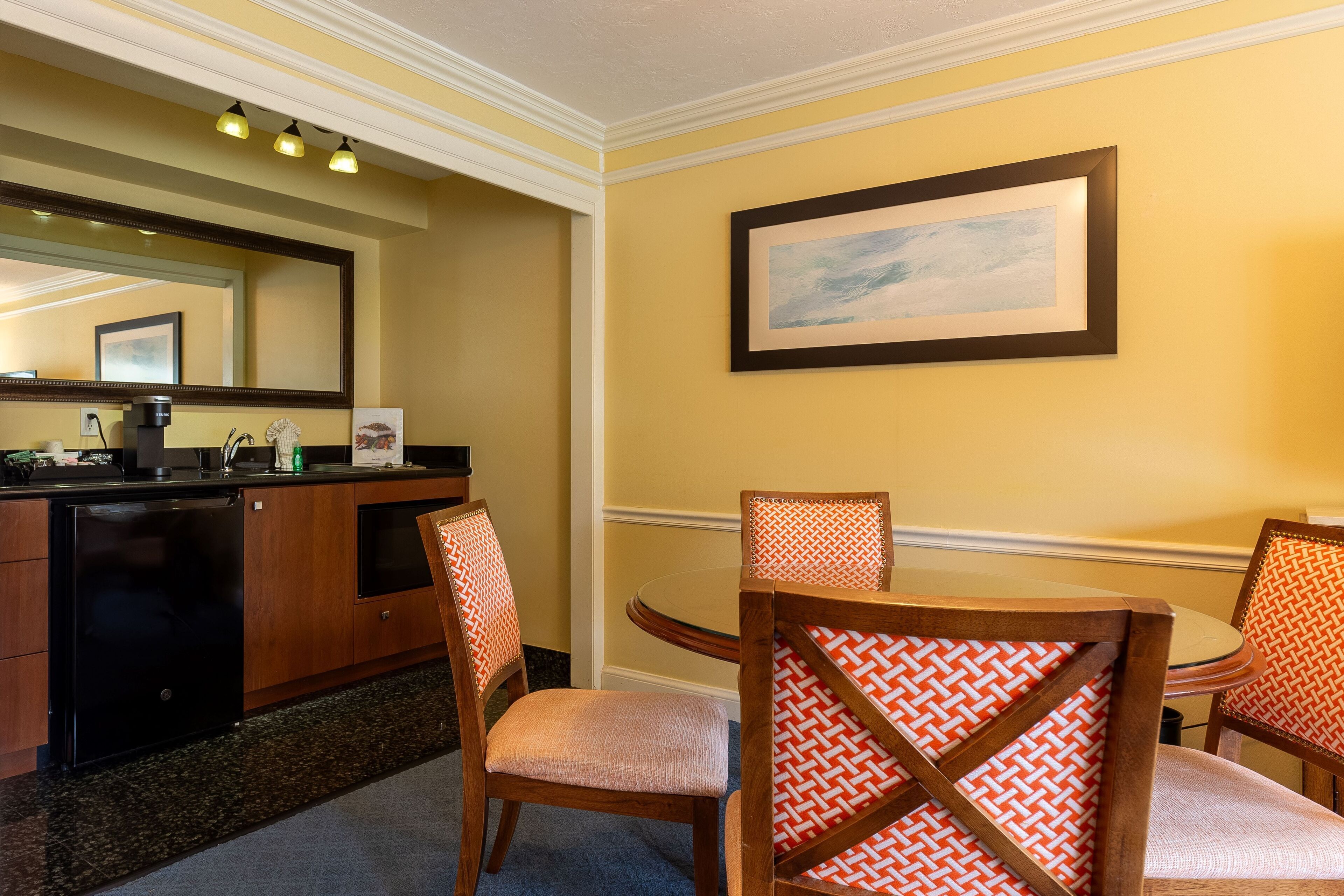 Une chambre d’hôtel dotée d’un coin repas comprenant des chaises en bois avec des coussins à motifs, une table ronde et une grande œuvre d’art encadrée au mur.