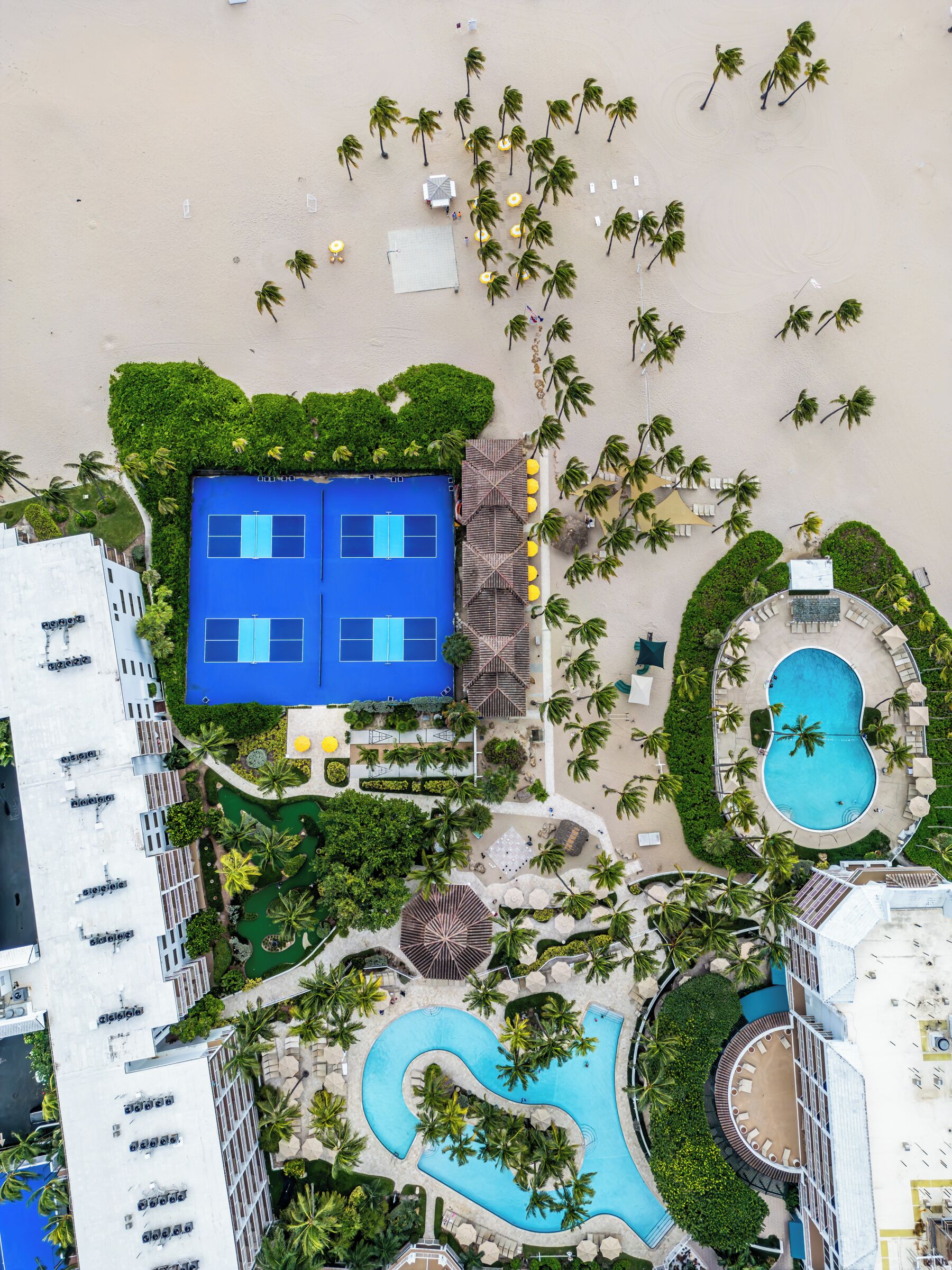 Ett flygbildsbild av ett semesterområde med en swimmingpool, tennisbanor och palmer.