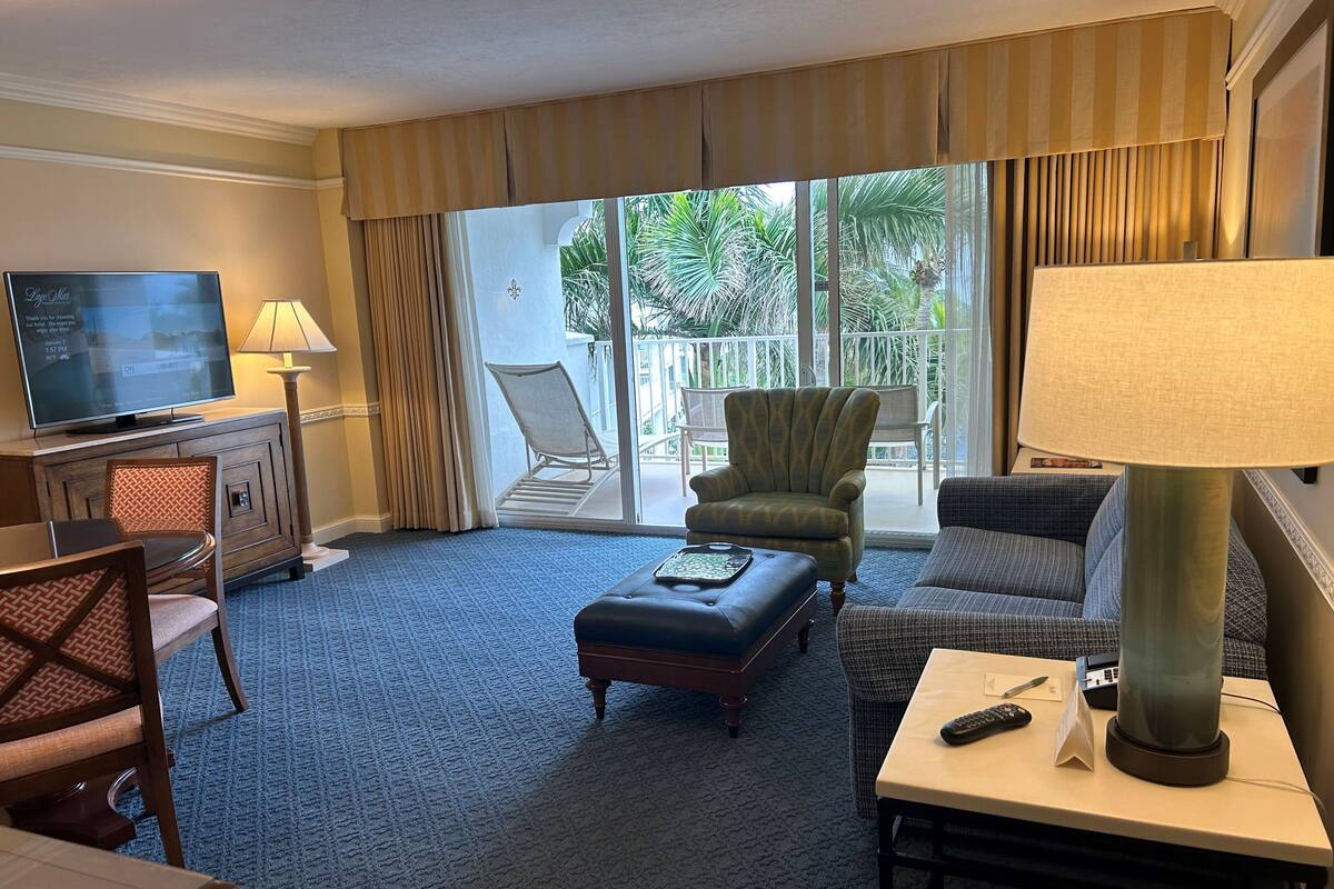 A hotel room with a television, a sofa, a chair, a table with a remote, and a balcony with a chair.