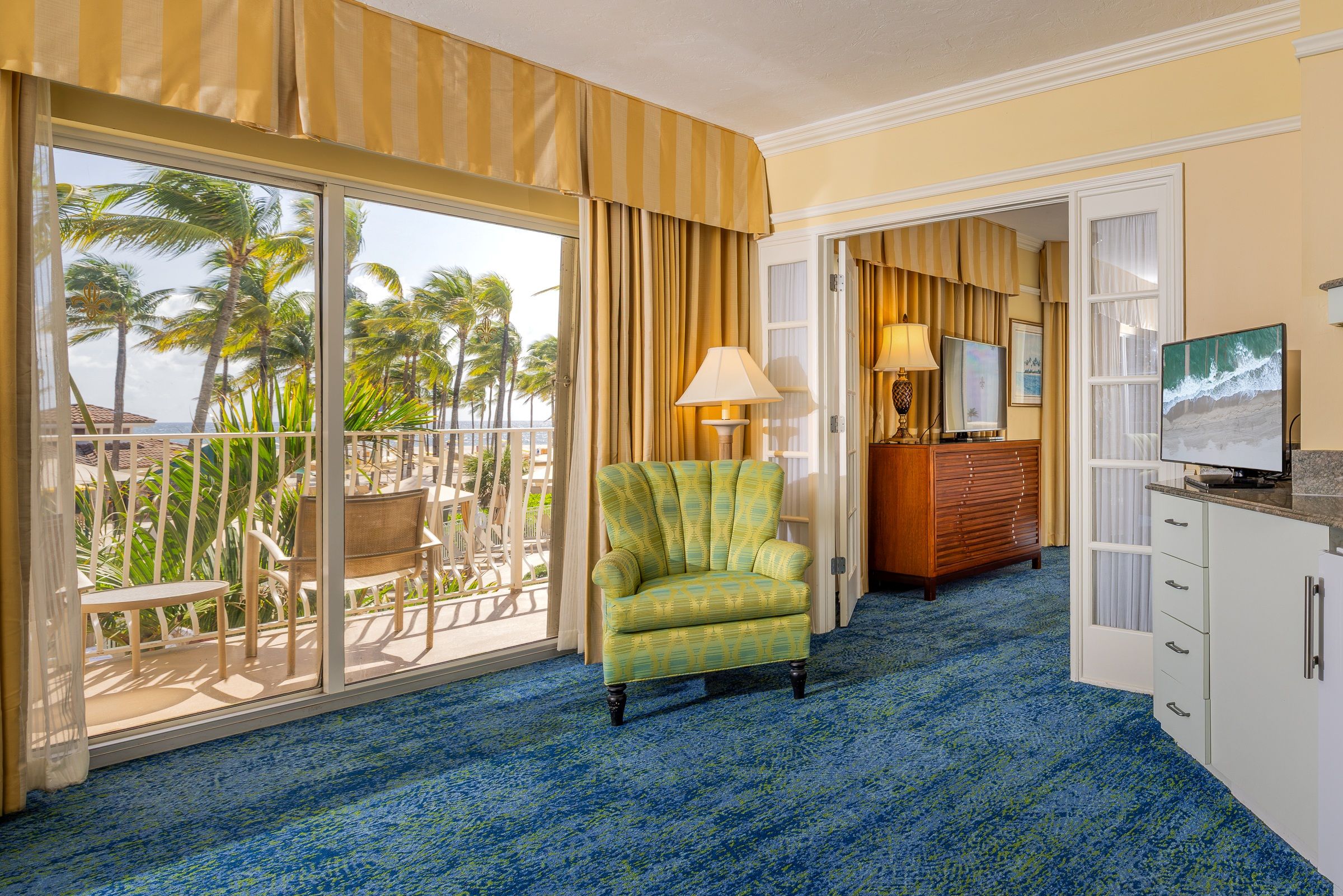 A hotel room with a balcony, a chair, a desk, a television, and a view of palm trees.