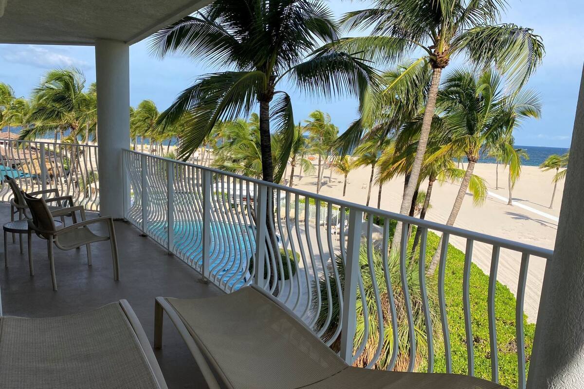 A balcony with lounge chairs overlooking a beach with palm trees and a pool.