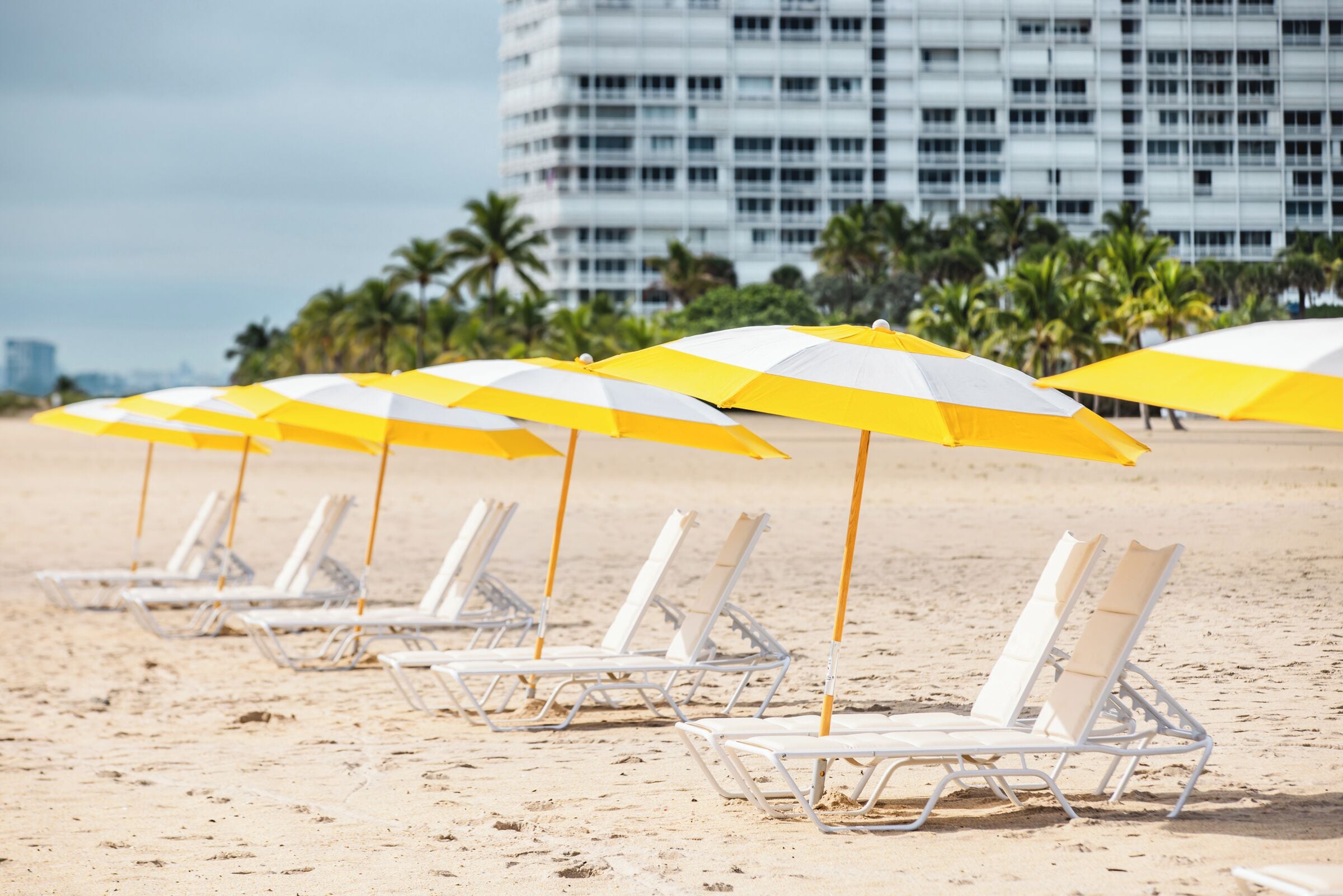 Een rij witte strandstoelen met gele en witte streepjes parasols op een zandstrand, met palmbomen en een modern gebouw op de achtergrond.